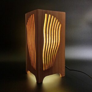Lampe artisanale en merisier massif allumée, lumière chaude diffusée par découpes ondulées