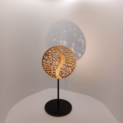Sculpture miniature en bois de noyer ajouré, disque circulaire aux motifs organiques évoquant racines ou canopée, montée sur tige métallique noire avec socle rond, projetant une ombre graphique sur le mur